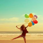 Een meisje springt op het strand met veel kleurige ballonnen. Op de achtergrond zee en blauwe lucht. 